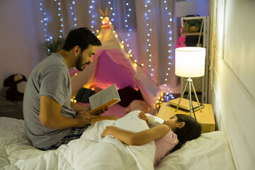 Father and young daughter enjoying a bedtime story in a cozy bedroom with a magical indoor tent and...