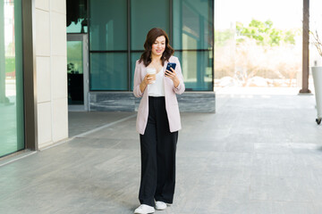 Self-assured female executive multitasking with smartphone and coffee while walking around the...