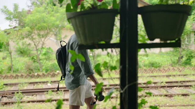 Asian traveler with backpack, camera strolls Haputale station. Male explorer in hat inspects rural train track. Solo journey, cultural visit in rich Sri Lanka. Authentic travel experience recorded.