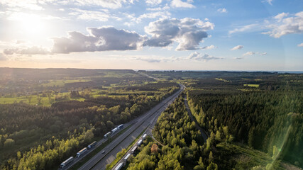 This aerial image captures the E42 highway as it winds through the verdant forests of the Hautes Fagnes region near sunset. The road is flanked by dense woodlands, showcasing a breathtaking landscape