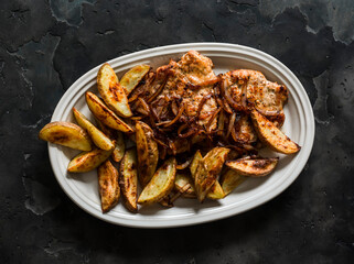 Delicious lunch, dinner - pork chop, baked new potatoes and caramelized onions in one plate on a dark background, top view