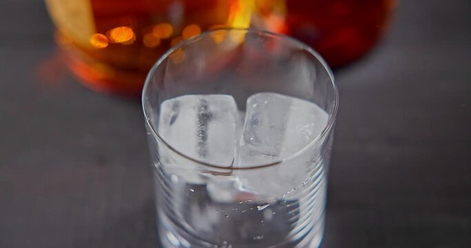 bartender puts cold ice cubes for cooling into a glass whiskey glass on a background of bottles