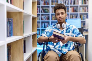 Front view portrait of teenage boy with disability using wheelchair in school library and holding...
