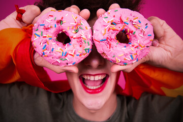 A funny guy dressed as a drag queen and eating pink donuts. Pink background.