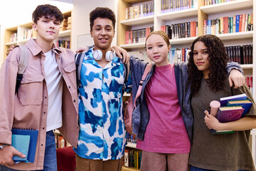 Waist up portrait of multiethnic group of students standing in school library holding books and smiling at camera