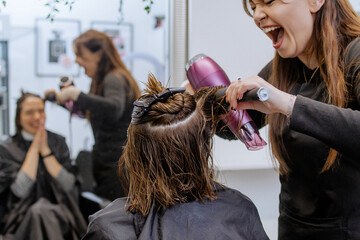 Positive, emotional woman professional hairdresser doing hair styling to client in beauty salon,...