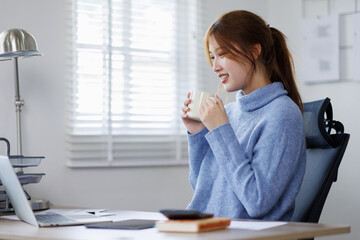 Freelancer asian woman working from home and using laptop sitting in living room with cup of hot...