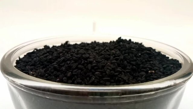 Black cumin seeds or black caraway seeds drops in a glass bowl on white background, a glass bowl full of black cumin seeds in the center, rotating, micro shot, nigella sativa seeds, herbs and spices