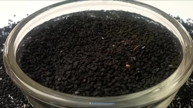 Black cumin seeds or black caraway seeds drops in a rotating glass bowl on white background with spices spilled around it, micro shot, center frame, herbs and spices