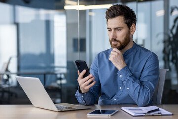 A serious and pensive young man is sitting in the office at a desk with a laptop and looking...