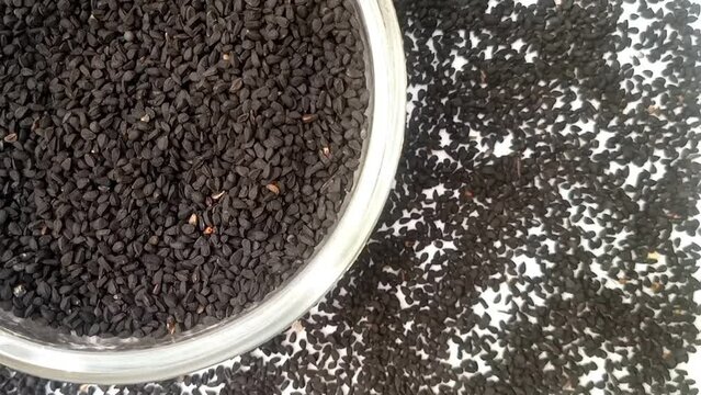 Black cumin seeds or black caraway seeds in a glass bowl rotating top left corner with spices spilled around it, white background, closeup, herbs and spices