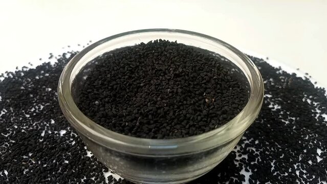 Black cumin seeds or black caraway seeds in a glass bowl spices spilled around it, rotating on white background, center frame, zooming in, close up, herbs and spices