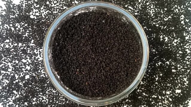 Black cumin seeds or black caraway seeds in a glass bowl spices spilled around it, rotating on white background, center frame, top view, close up, herbs and spices