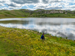 girl picking field yellow flowers on the river bank with reflections of clouds in the water and varatic rocks in the background