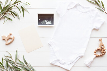 Baby Bodysuit mockup. Blank white baby bodysuit on a wood background with card, toys and ultrasound photo, flat lay. Pregnancy Announcement template, copy space.