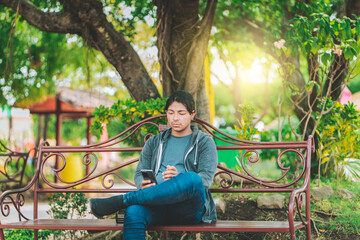 Handsome man sitting on a bench checking cell phone at sunset. Young man sitting on a park bench texting on cell phone