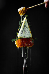 A slice of blue cheese, served with honeycomb and honey on a fork. On a black background.