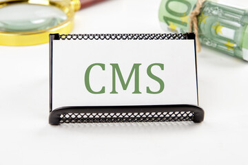 CMS (Custom Management System) written on a white card on a white background in a composition with EURO bills and a magnifying glass