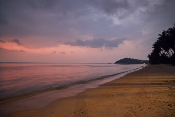 Evening as the sun sets over a calm sea, with a sandy shore lined by silhouetted palms