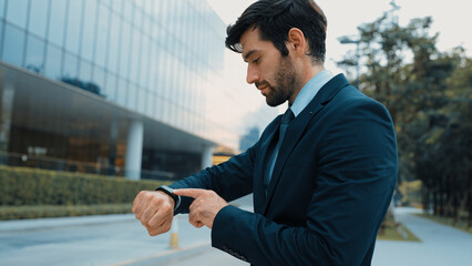 Skilled caucasian business man looking at time while standing at building. Attractive executive...