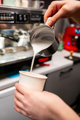 Hands pouring milk from a stainless steel pitcher into a paper cup, with kitchen appliances in the...