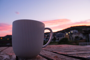 As the sun sets, a coffee cup is placed on a wooden table against a backdrop of the colorful sky....