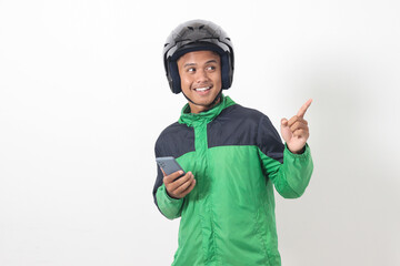 Portrait of Asian online taxi driver wearing green jacket and helmet holding mobile phone while pointing to the side. Isolated image on white background