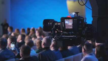 A detailed view of a professional video camera capturing a live event with a crowd of attendees in a conference hall. Focus on technology and event documentation.