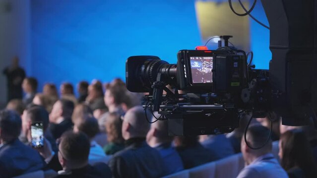 A detailed view of a professional video camera capturing a live event with a crowd of attendees in a conference hall. Focus on technology and event documentation.