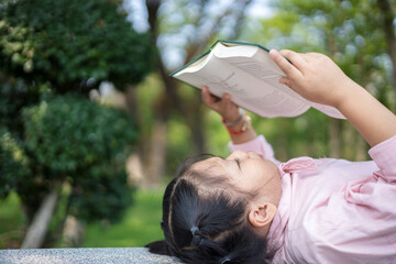 Cute little asian girl reading a book while lying at garden