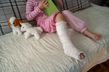 Little girl with broken leg, reading a book playing with a toys, unrecognizable . Leg in plaster...