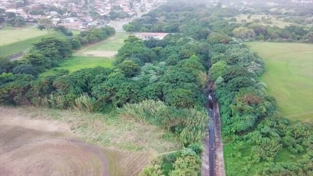 drone flying over a watery sewerage canal surrounded by bush and sports fields abandon shacks