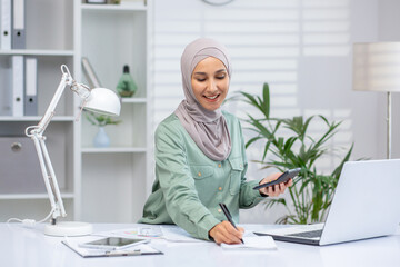 Smiling Muslim woman in hijab seated at a modern office, using a smartphone and laptop. She...