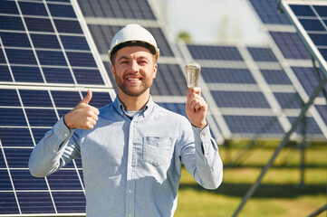 Smiling and holding bulb. Engineer with photovoltaic solar panels outdoors at daytime