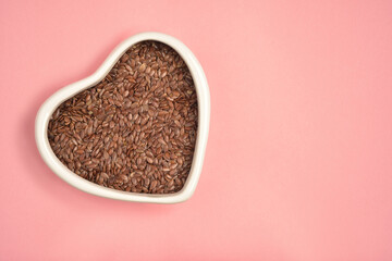 Photograph of brown flaxseed or linseed in heart shape white bowl with copy space on pink colour background. Top view. Health care concept.