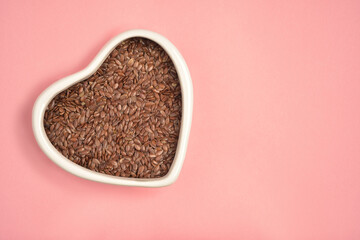 Photograph of brown flaxseed or linseed in heart shape white bowl with copy space on pink colour...