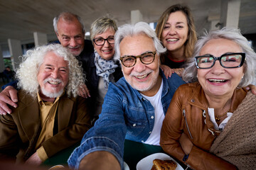 Happy mature gray hair man taking selfie of group seniors Caucasian cheerful friends posing together sitting at cafeteria. Older people looking smiling at camera with snacks on table nursing home