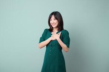 Adorable happy young asian woman model with shy emotion smiling expression cheerful positive attractive face and holding hand isolated on green pastel studio background. Portrait beautiful cute lady.
