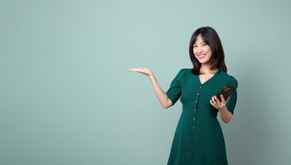 Asian woman green dress gesturing hand open to free copy space while holding mobile phone isolated on pastel green background. online payment, promotion, deals concept.