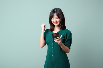Asian woman in a green dress is receiving good news from mobile phone against a green background.