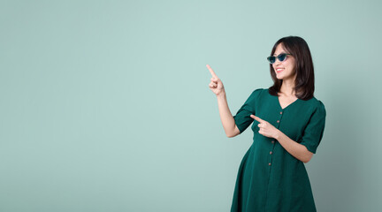 Asian woman with sunglasses and green dress pointing finger to free copy space isolated on pastel green background. promotion, deals concept.