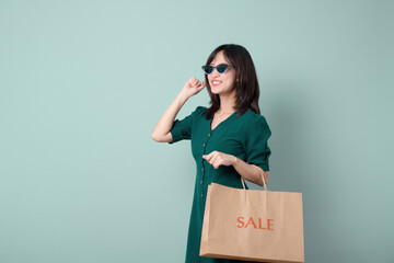 Young beautiful Asian woman with sunglasses and green dress smiling and holding paper shopping bag isolated on green background, Supermarket and fashion mall concept