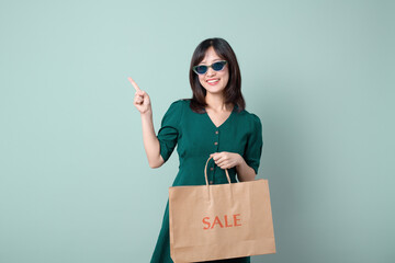 Young beautiful Asian woman with sunglasses and green dress smiling and holding paper shopping bag isolated on green background, Supermarket and fashion mall concept