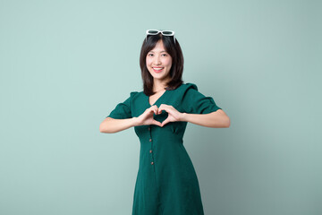Asian woman green dress posing hand heart gesture isolated on green studio background. Healthcare concept.