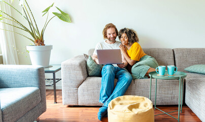 Multiracial young couple watching computer laptop sitting on the sofa at home - Happy diverse...