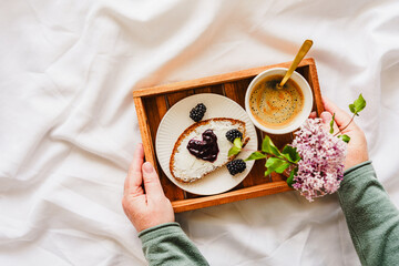 Eine Frau hält ein Holz Serviertablett mit einer Tasse Kaffee, süssen Frühstück und Blumen auf einem Bett. Zuhause.