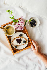 Ein weibliche Hand hält ein Serviertablett mit einer Tassee Kaffee, ein Marmeladenbrot in Herzform und Blumen auf einer weißen Bettdecke. Zuhause.