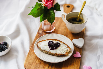 Eine Tasse Kaffee, eine Brotscheibe mit Konfitüre in Herzform und eine Rose auf einem Holz...
