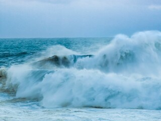 blue ocean wave splashes in front of small lighthouse with people watching