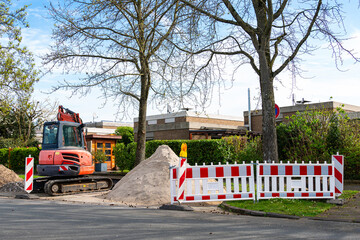 A small crawler excavator repairs a section of road. The repair site is fenced with red and white...